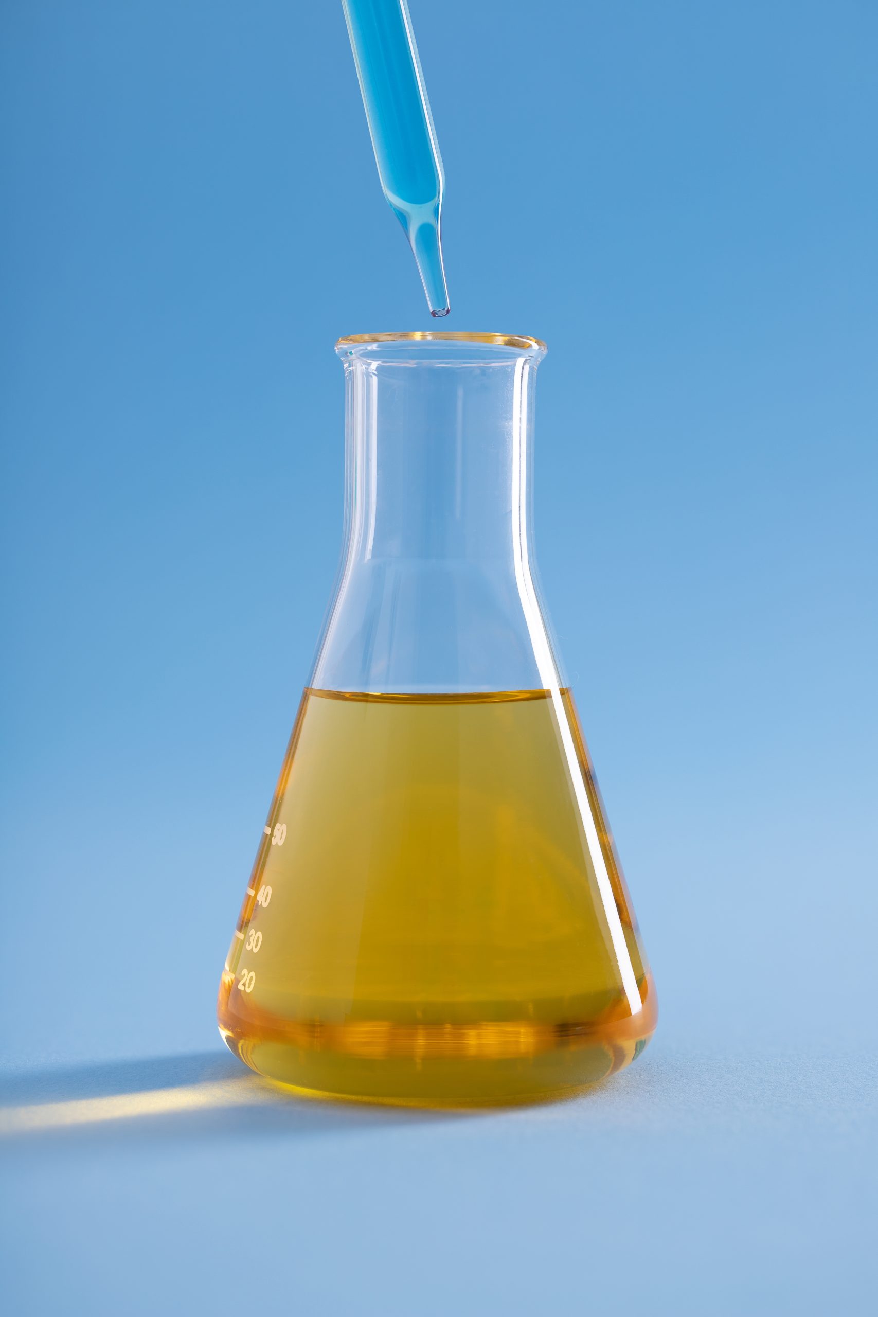 A vertical closeup of a dropper with blue liquid over an Erlenmeyer flask with yellow liquid on a blue background